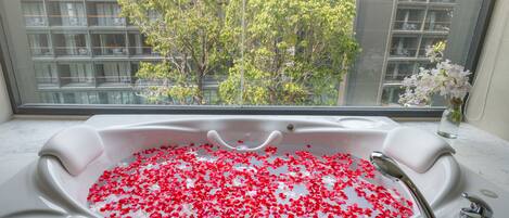 Una bañera llena de agua y pétalos de rosa, con vista a un árbol y edificios a través de una ventana.