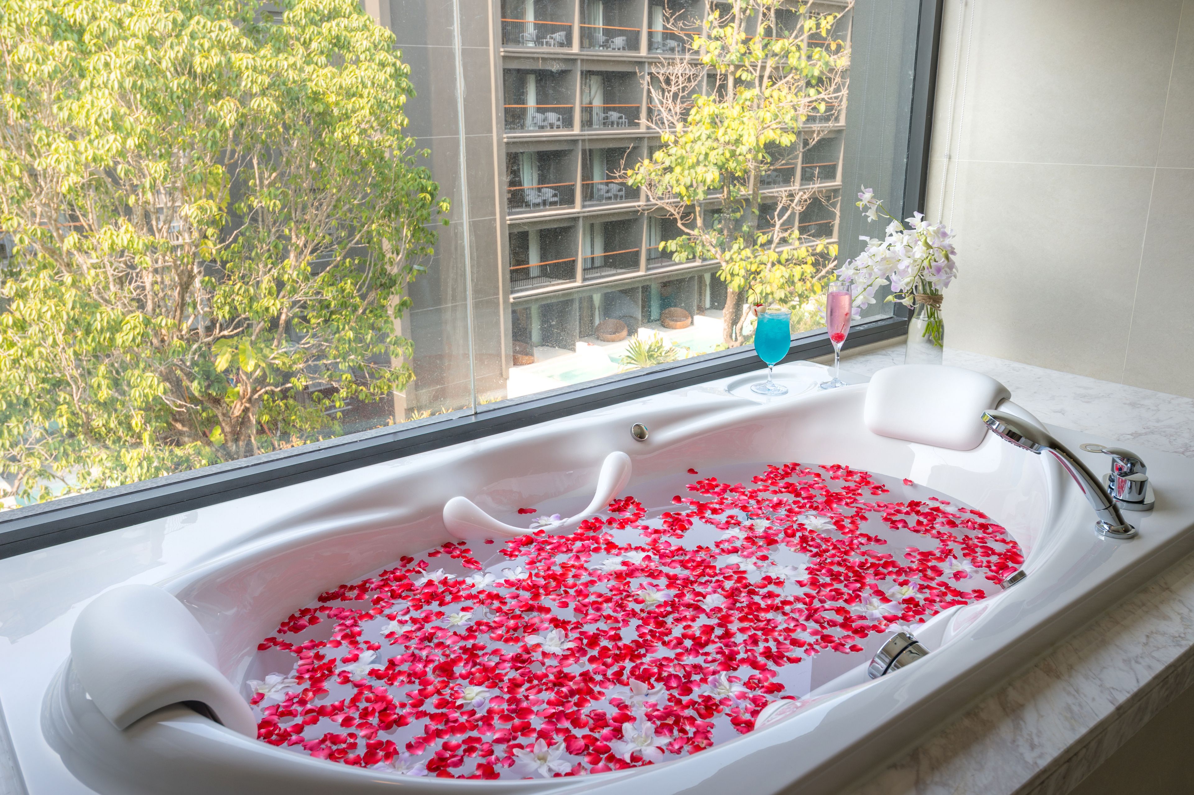 A bathtub filled with rose petals and a glass of blue drink, with a view of trees and a building outside.