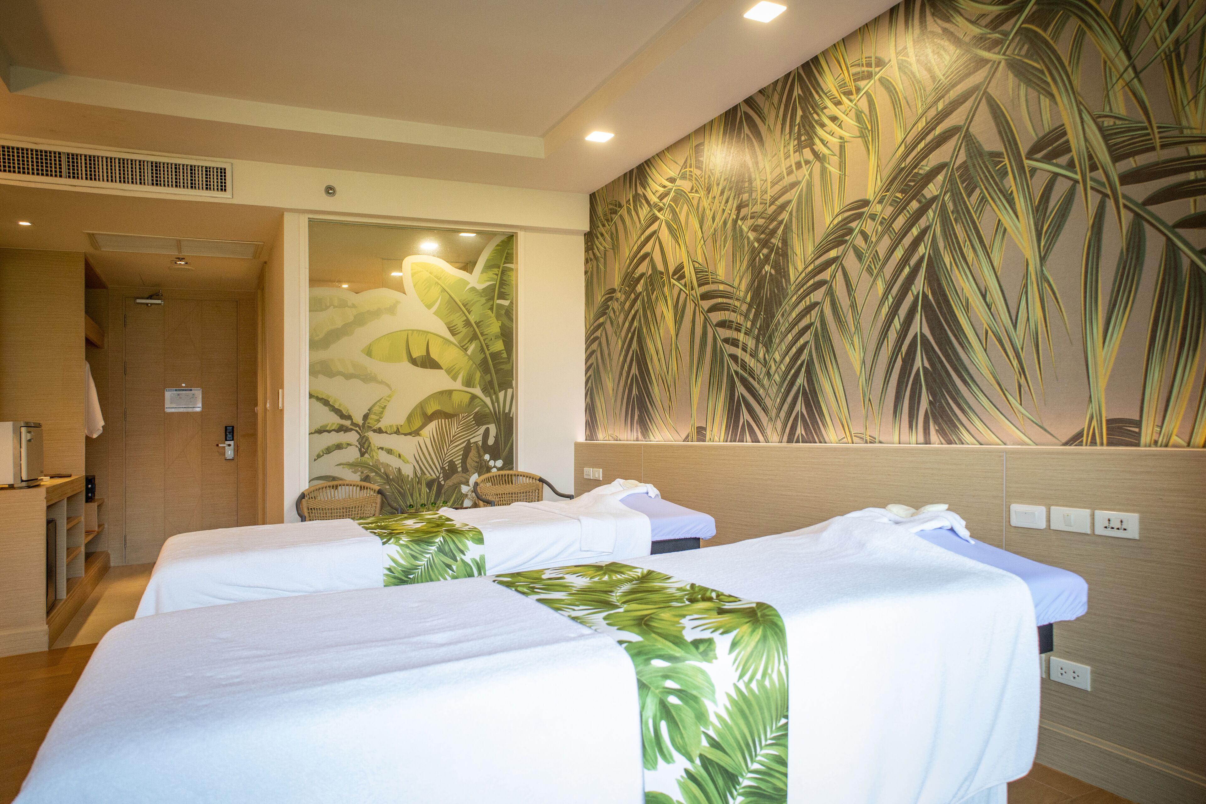 A hotel room with two beds, a large tropical-themed mural on the wall, a wooden door, and a small seating area with a wicker chair.