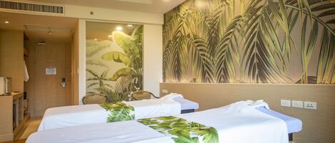 A hotel room with two beds, a large tropical-themed mural on the wall, a wooden door, and a small seating area with a wicker chair.