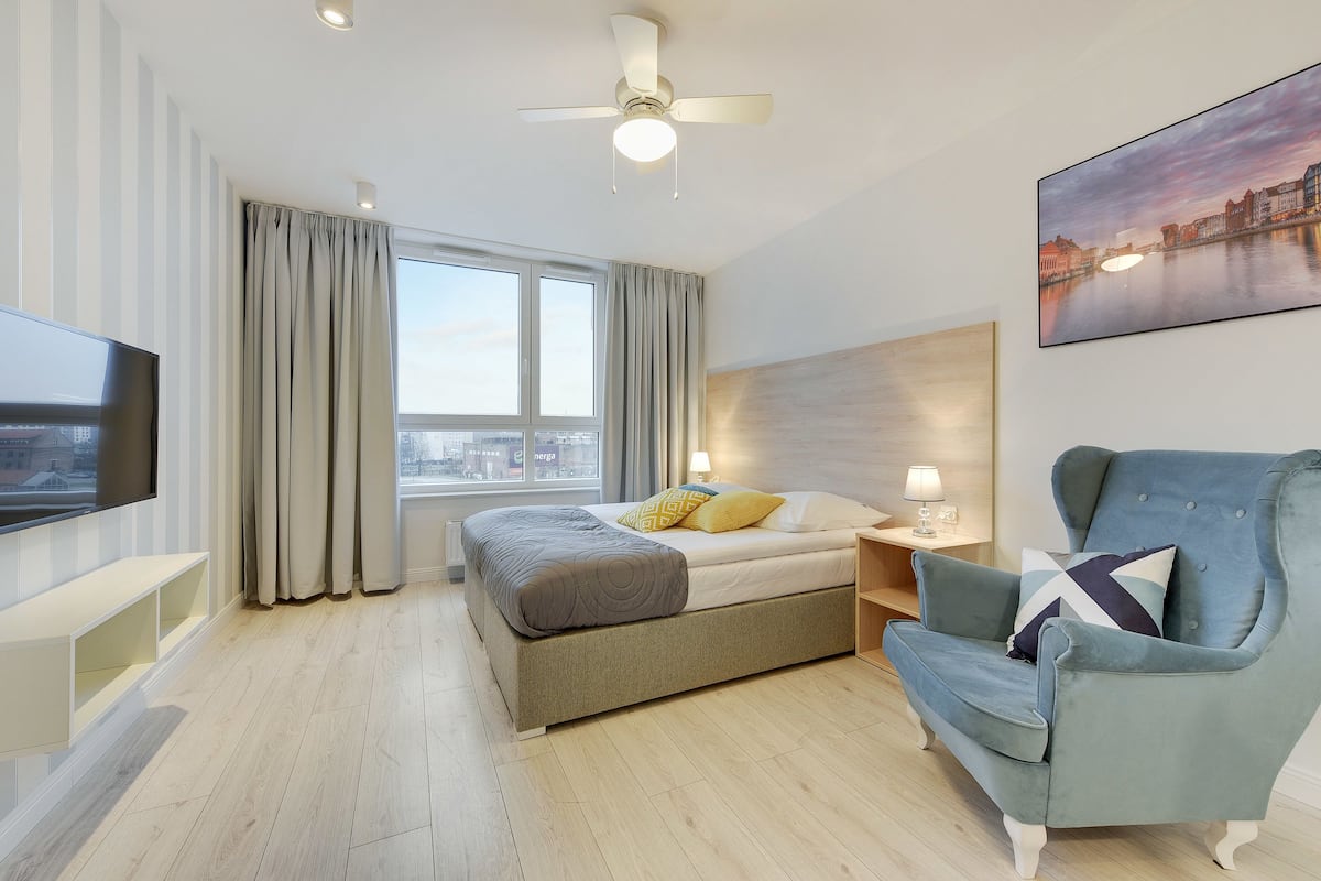 Une chambre d’hôtel moderne équipée d’un lit, d’une télévision, d’une chaise, d’une fenêtre avec des rideaux et d’un ventilateur de plafond.