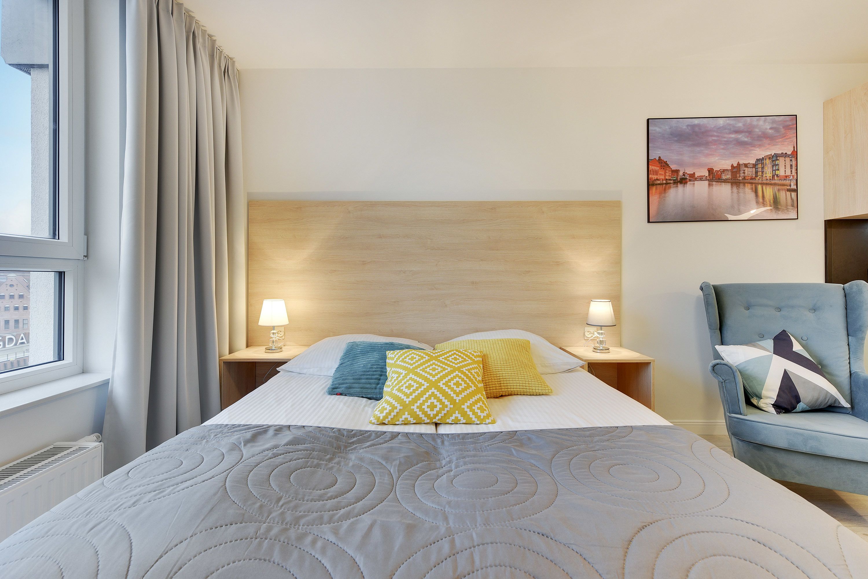 Habitación de hotel con una cama grande, dos lámparas de noche, un sillón azul y un cuadro con una vista urbana.