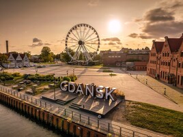 Una zona ribereña con noria, un cartel grande que dice "GDANSK", y edificios históricos.