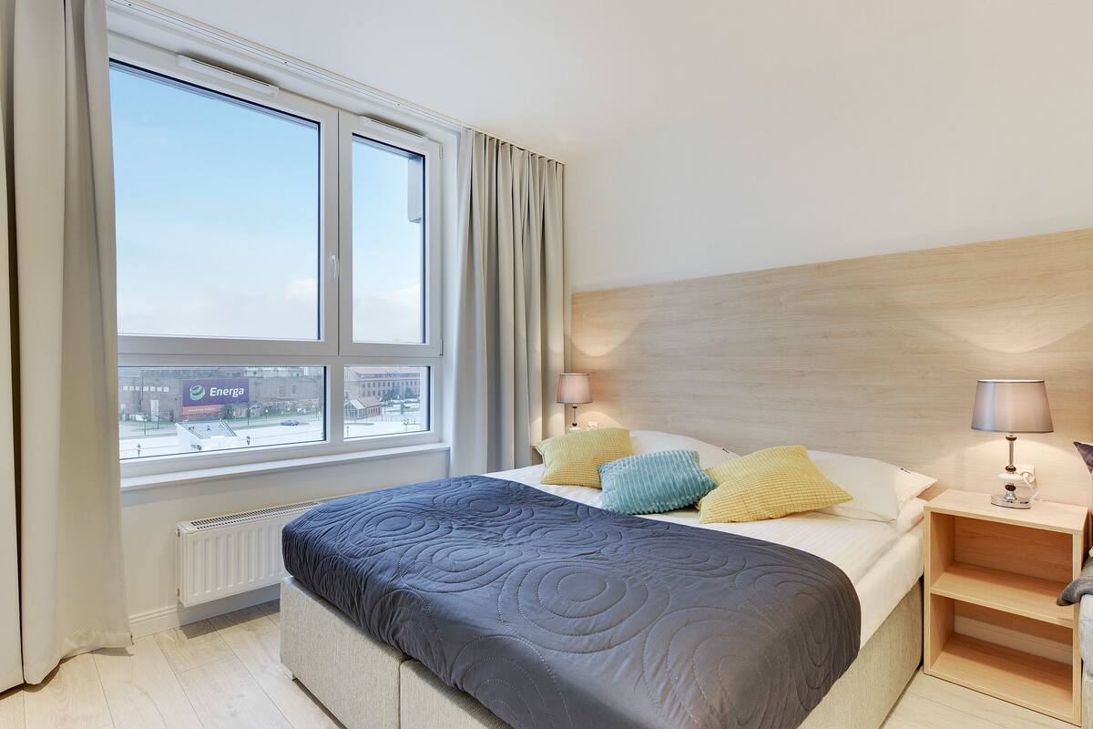 Une chambre d’hôtel avec un grand lit, une table de chevet avec des lampes, une vue sur la ville par la fenêtre et un radiateur placé sous la fenêtre.