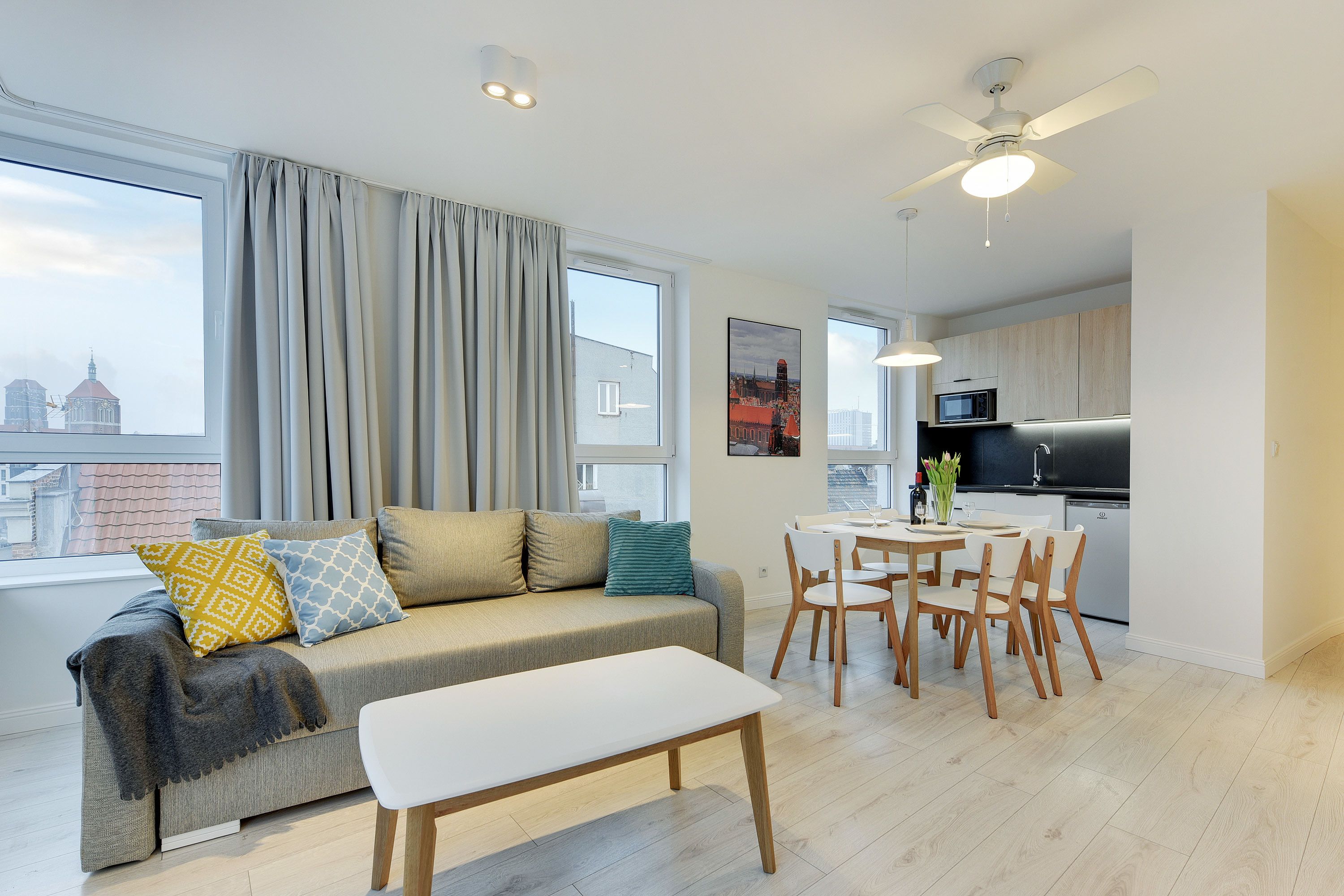 A modern living room with a sofa, a dining table, and chairs. There is a kitchen area with a microwave and a sink.