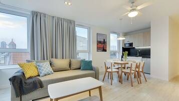 A modern living room with a sofa, a dining table, and chairs. There is a kitchen area with a microwave and a sink.