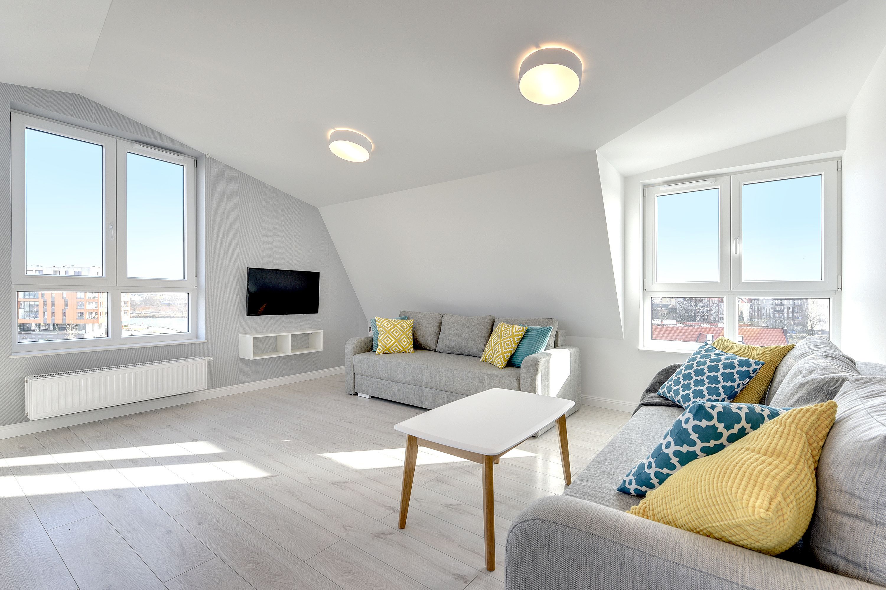 A modern living room with a grey sofa, a white coffee table, and patterned cushions. There are two large windows providing natural light and a view of the city.