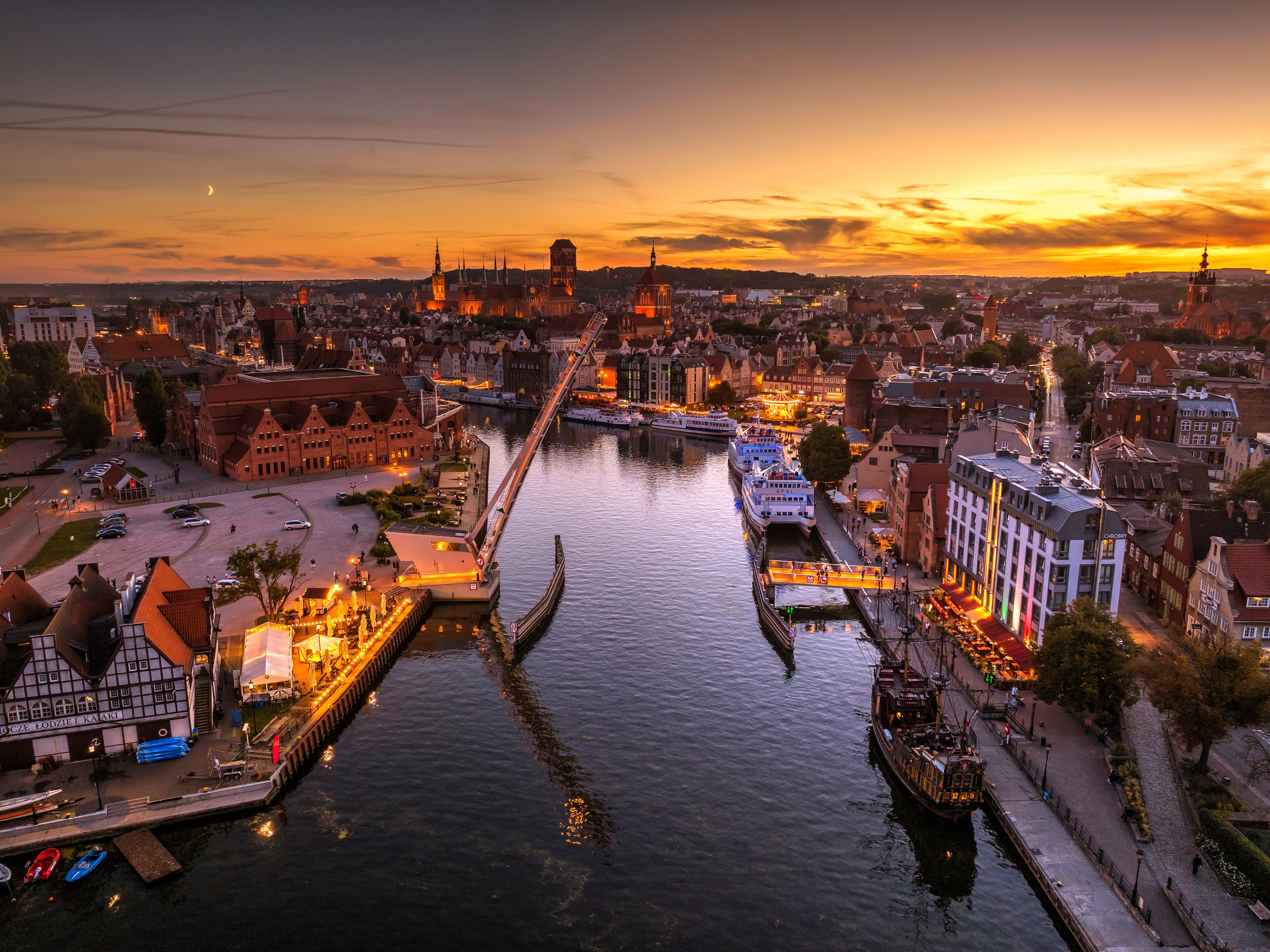 Une vue urbaine le long d’une rivière, avec des bâtiments d’époque, un pont et des bateaux amarrés au bord de l’eau.