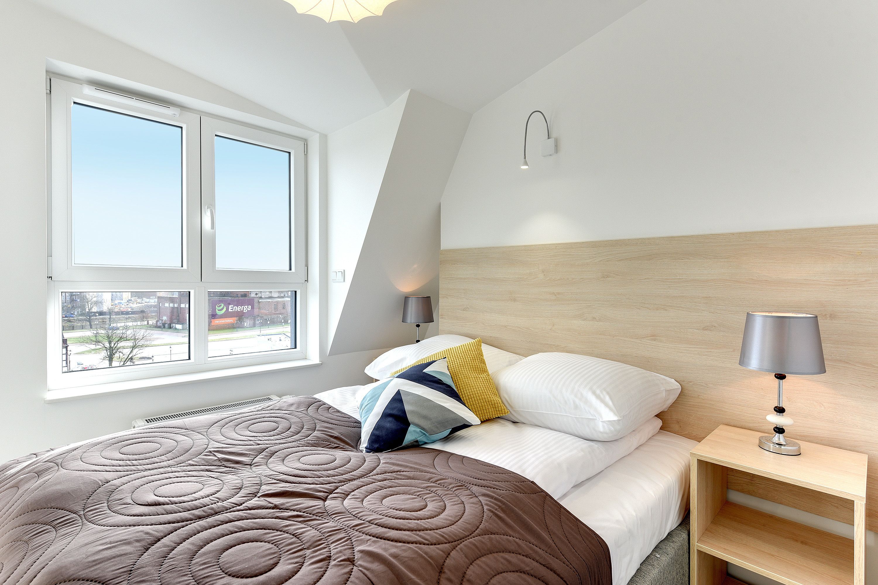 A modern bedroom with a large window, a wooden headboard, a bedside table with a lamp, and a quilted bedspread.