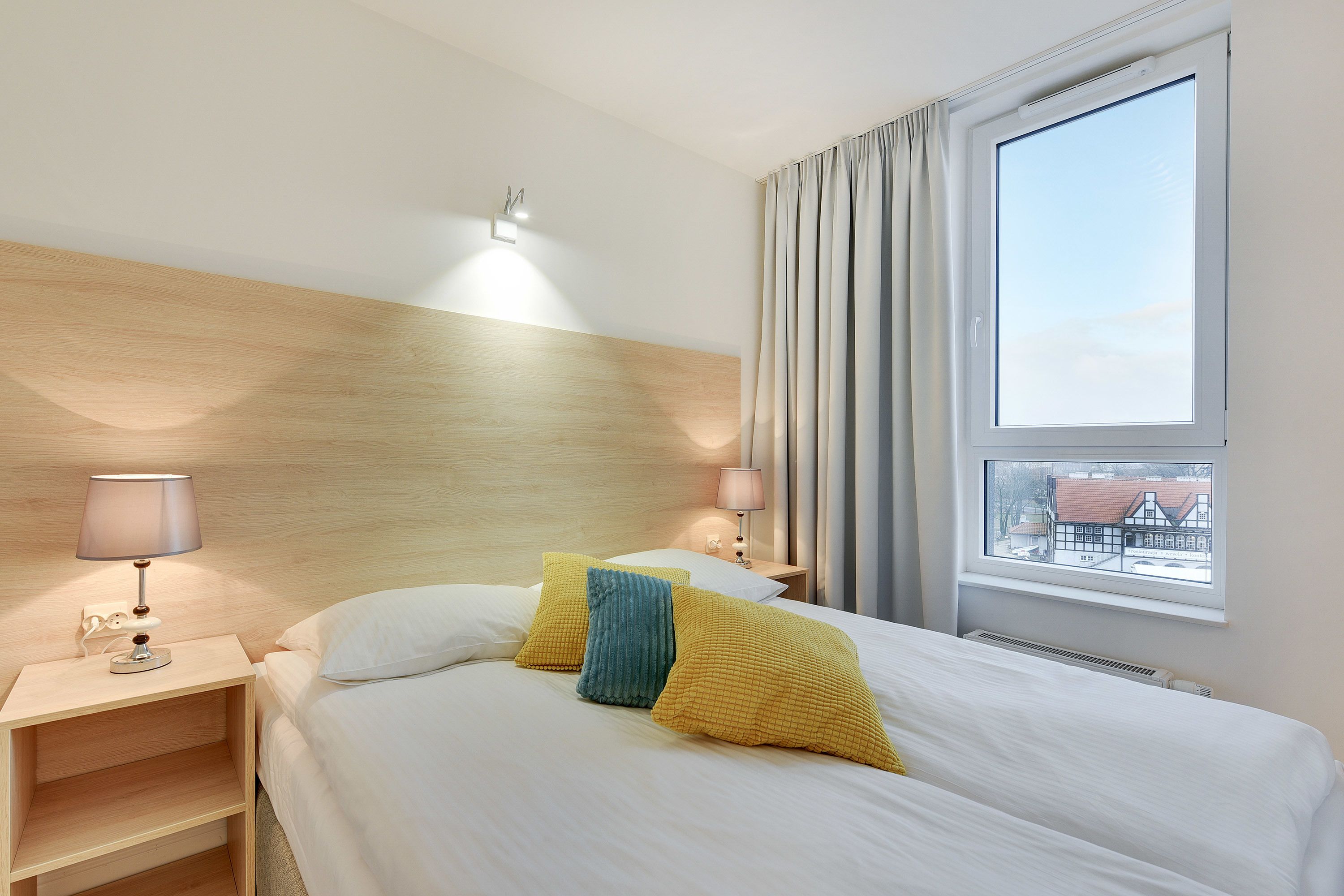 A hotel room with a wooden headboard, a bed with white bedding, a nightstand with a lamp, and a window with curtains.