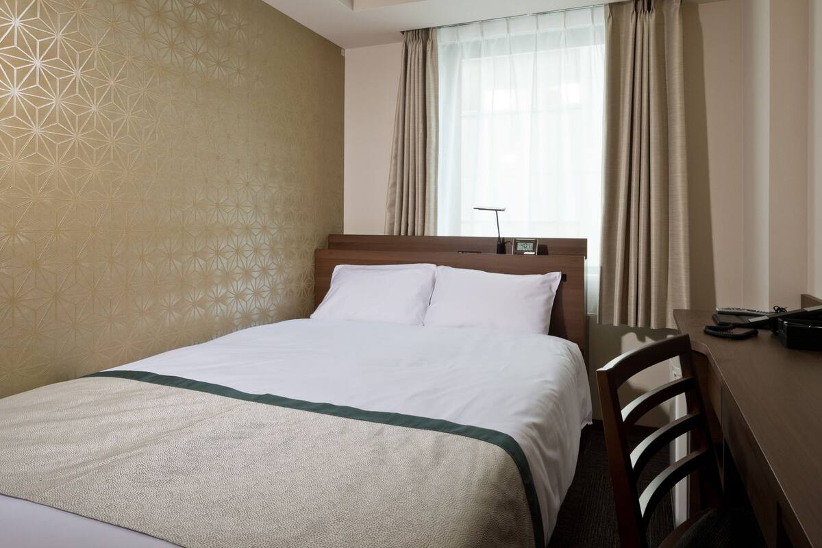 Een hotelkamer met een net opgemaakt bed, een bureau met stoel en een raam met gordijnen.