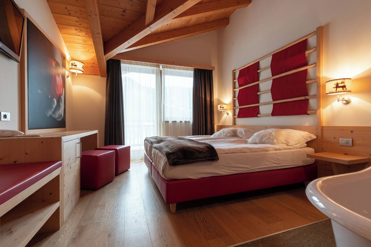 A modern hotel room with a wooden ceiling, a large bed with red bedding, a desk with a chair, a large window with curtains, and wall-mounted lights.