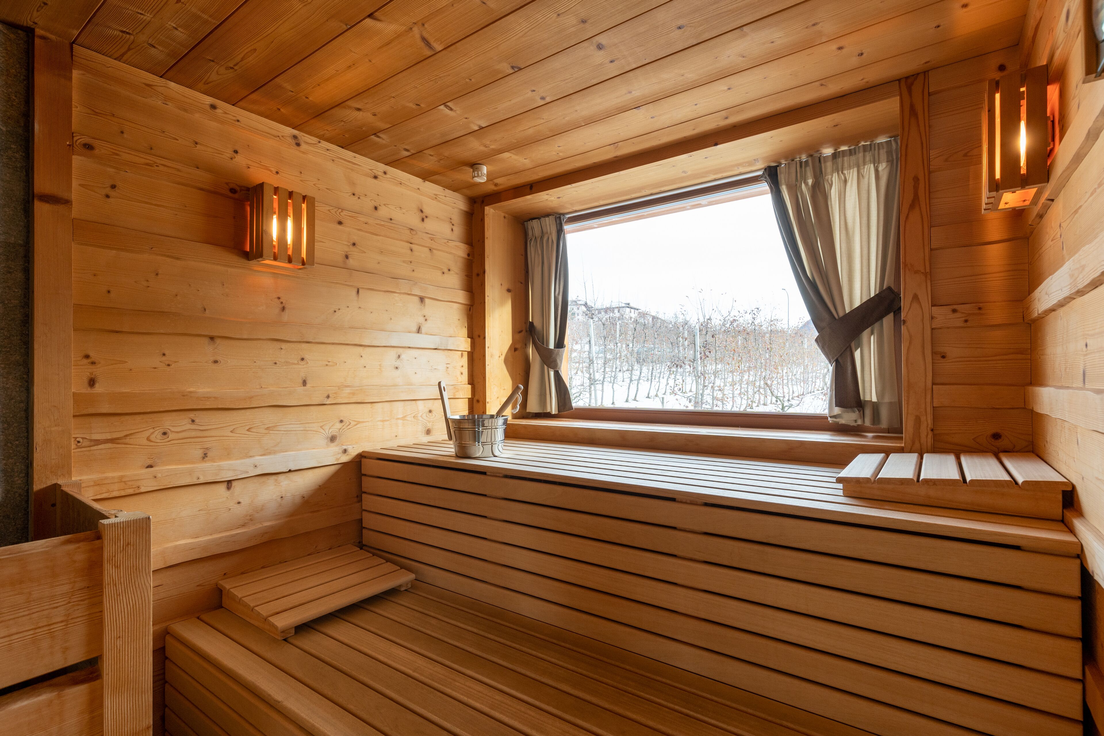 Interior de una cabaña de madera con un banco, una ventana con cortinas y una mesita con un hervidor.
