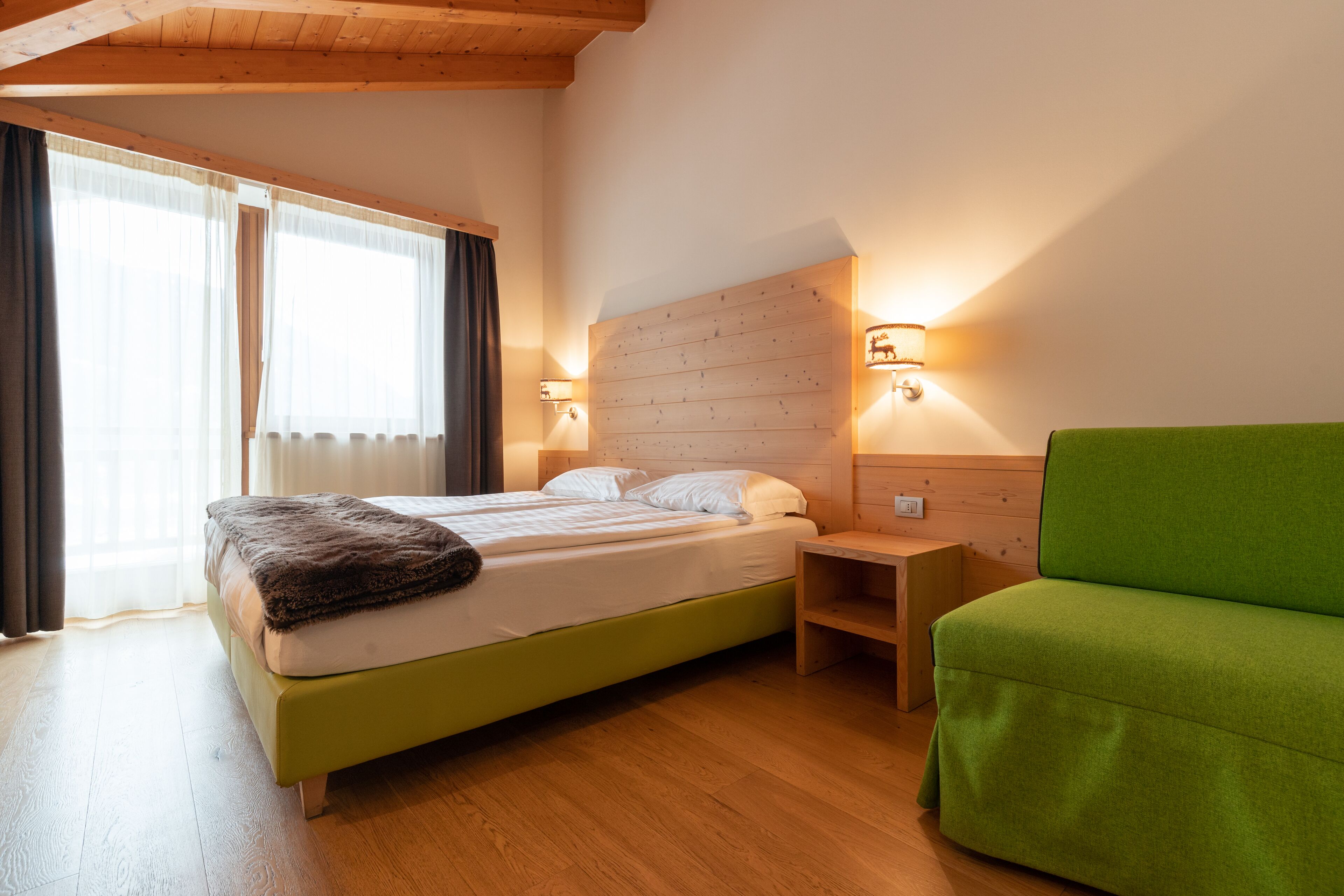 Una camera da letto con un letto, comodini, un divano verde, pareti in legno e una finestra con tende.