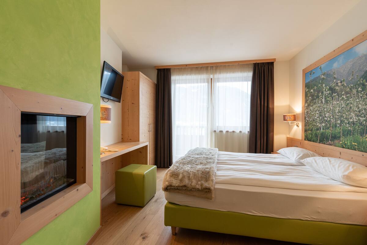 A hotel room with a bed, a television mounted on the wall, a window with curtains, and a painting on the wall.