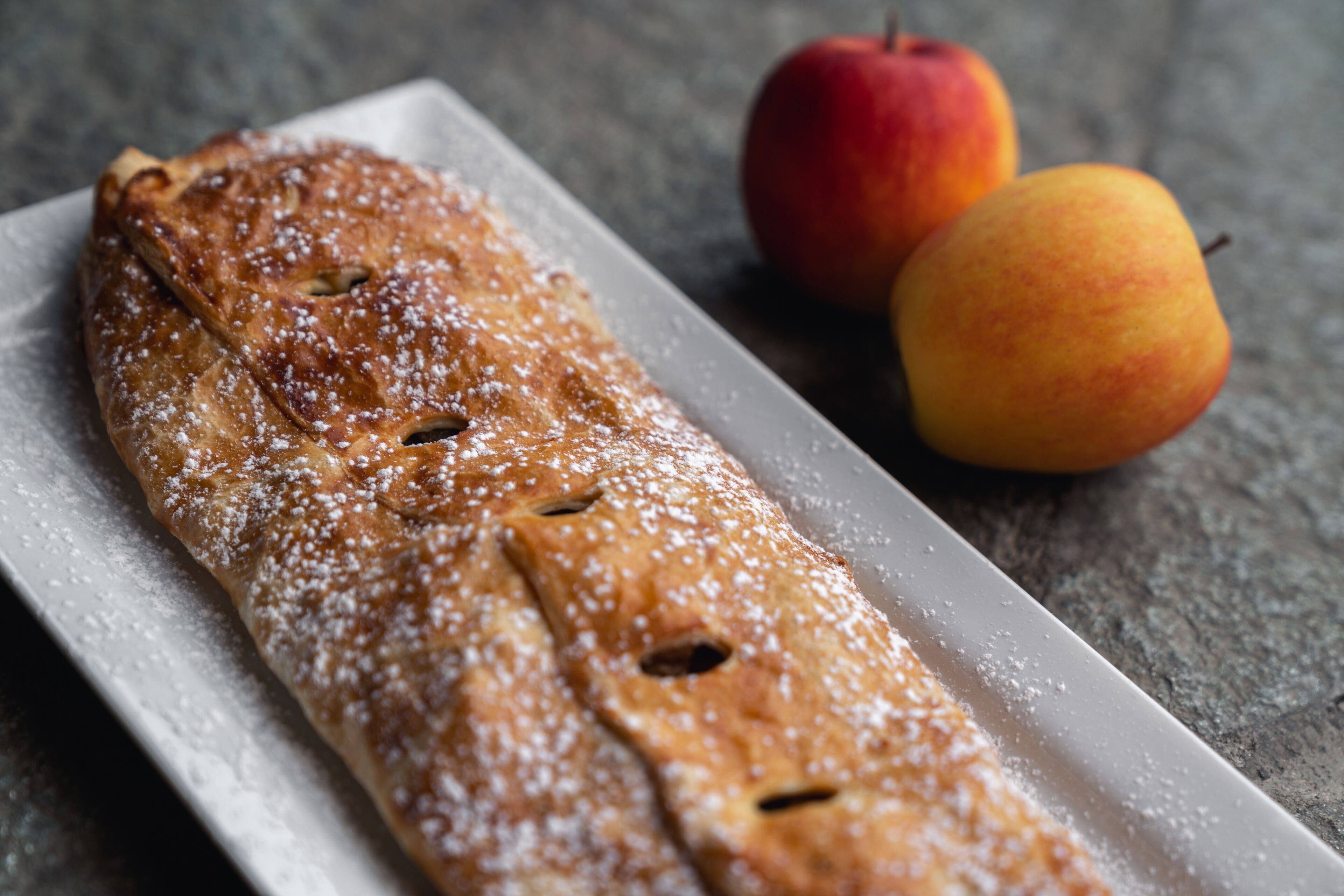 Een lang, rechthoekig gebakje bestrooid met poedersuiker, geserveerd met twee stukken fruit op een bord.