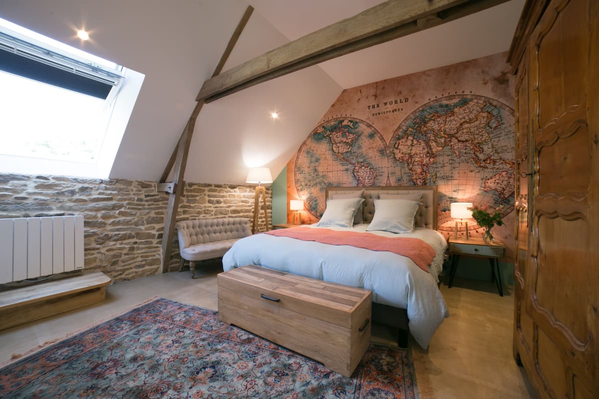 Un dormitorio con techo inclinado, un gran mapa del mundo en la pared, una cama con cabecera de madera, una mesita de noche con lámpara y un aparador.