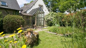 Uma casa de pedra com uma estufa de vidro, rodeada por um jardim bem cuidado com diversas plantas e flores.