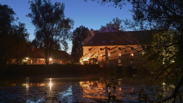 Un bâtiment en pierre doté de nombreuses fenêtres et dont l’extérieur est illuminé, situé au bord d’une étendue d’eau où se reflètent les lumières.