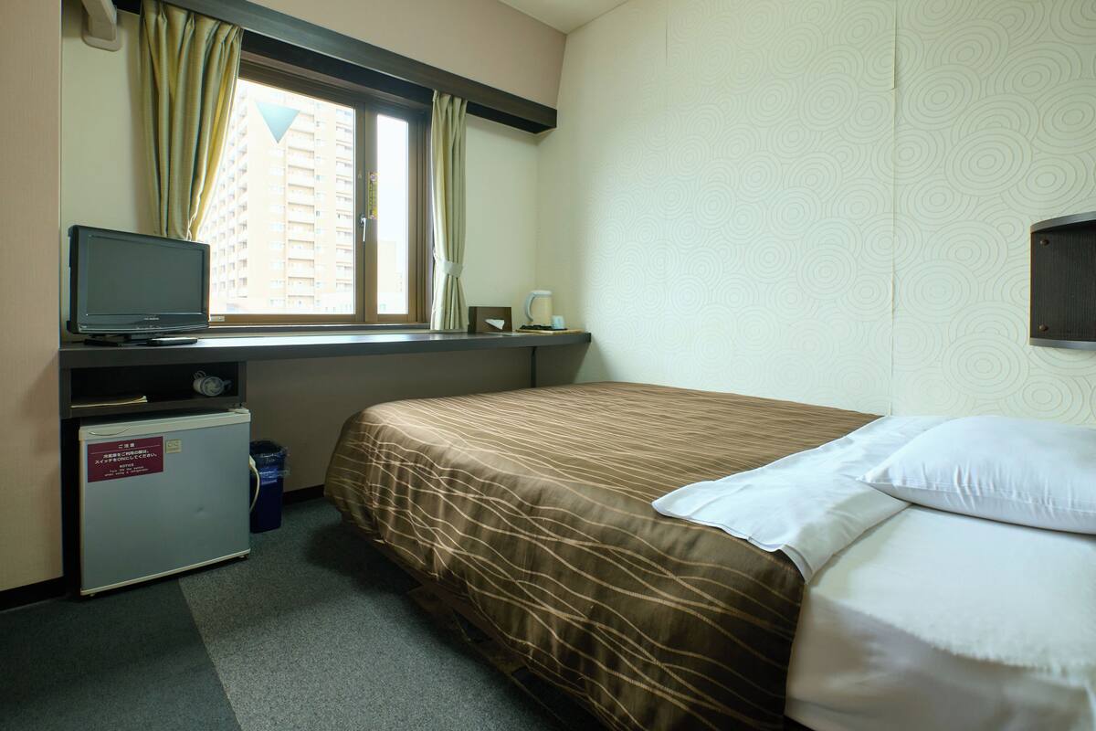 Hotellihuone, jossa on sänky, työpöytä, jossa on televisio, pieni jääkaappi ja ikkuna, jossa on verhot.