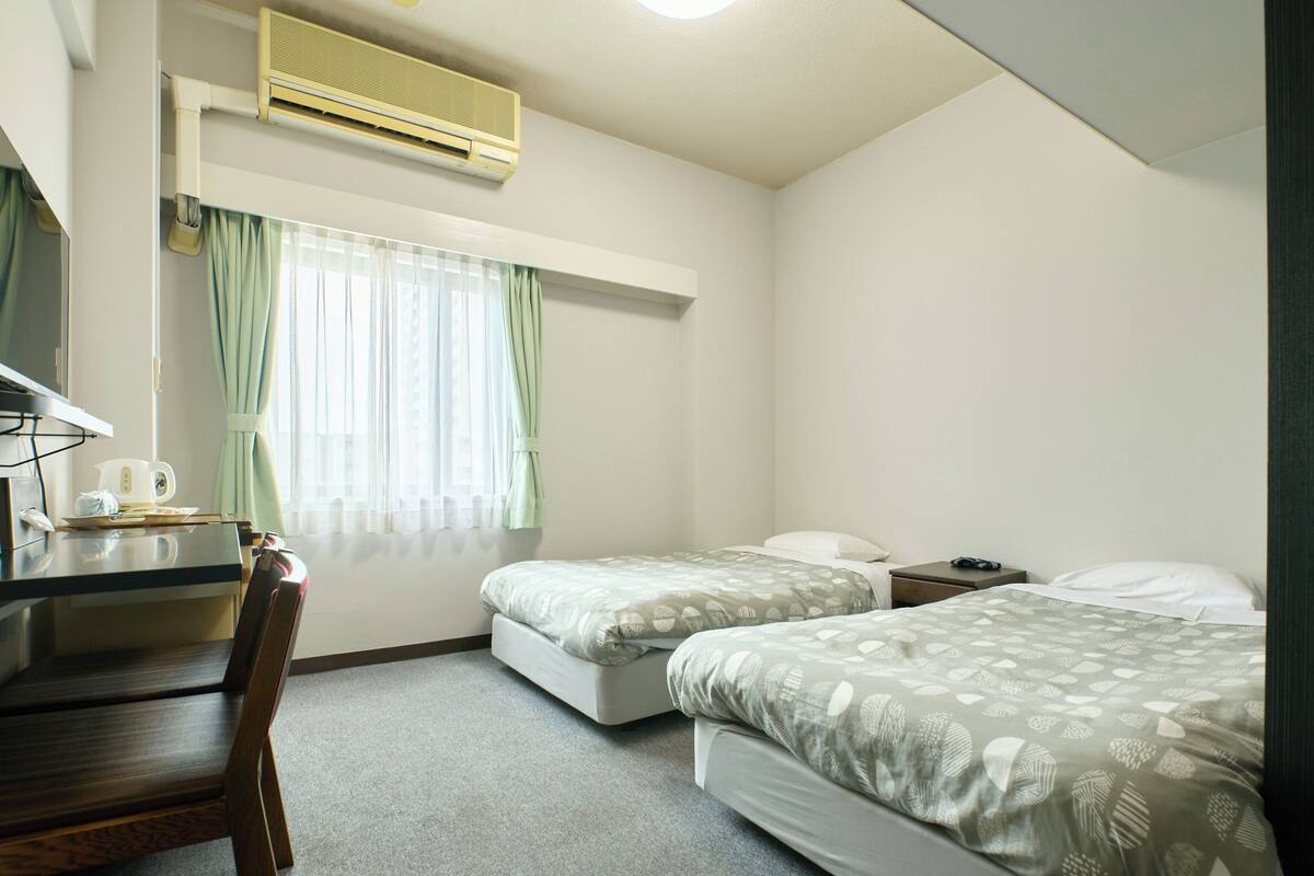 Hotellihuone, jossa on kaksi sänkyä, työpöytä, tuoli, pieni pöytä ja ilmastointilaite.