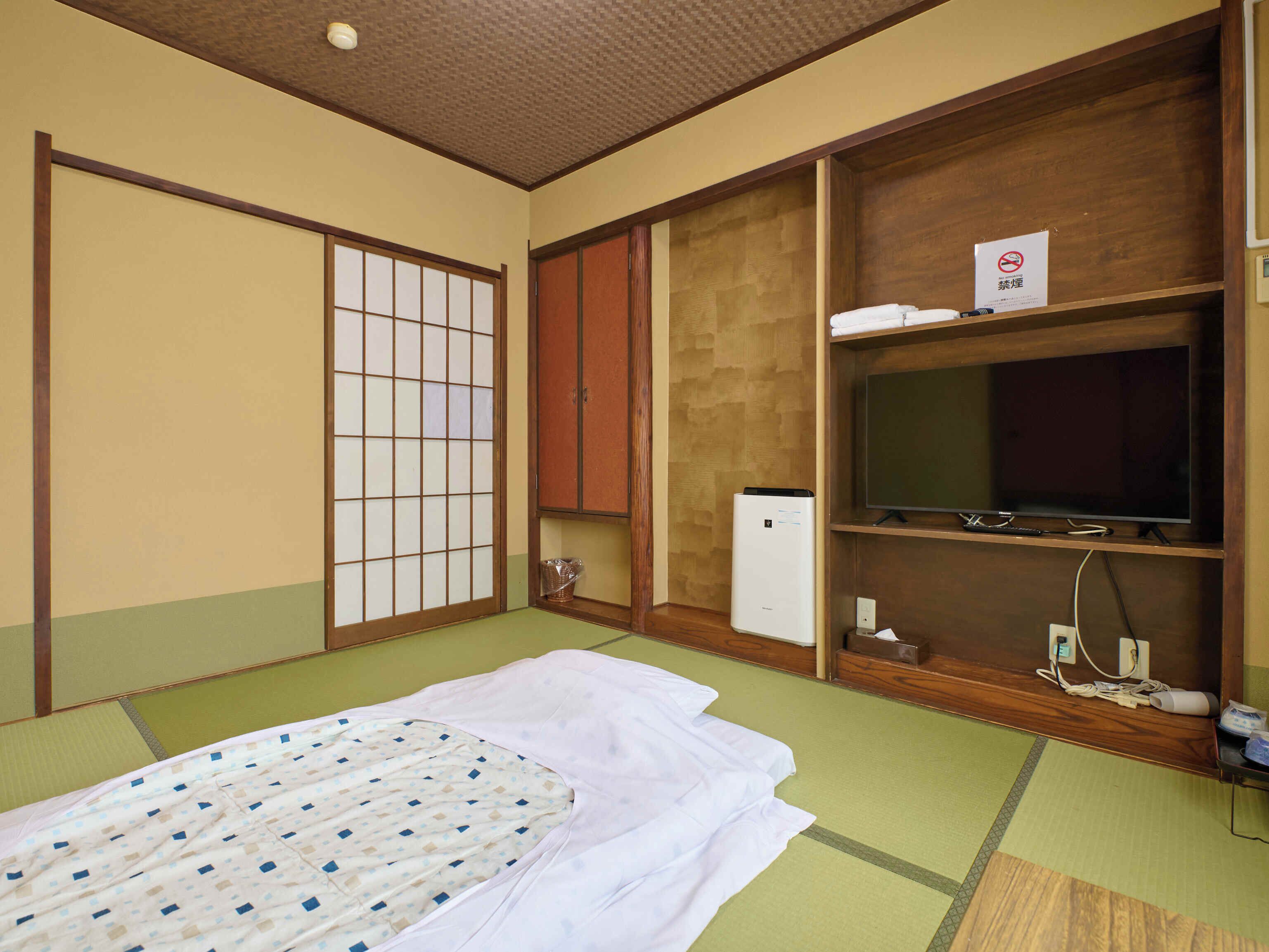 Una habitación japonesa tradicional con suelo de tatami, futón, puertas correderas, televisor y un refrigerador pequeño.