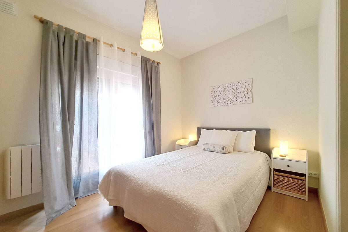 Ein Schlafzimmer mit einem großen Bett, zwei Nachttischen mit Lampen, einem Heizkörper und einem Fenster mit Vorhängen.
