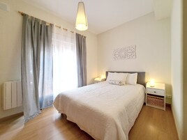 Un dormitorio con una cama grande, dos mesitas de noche con lámparas, un radiador y una ventana con cortinas.