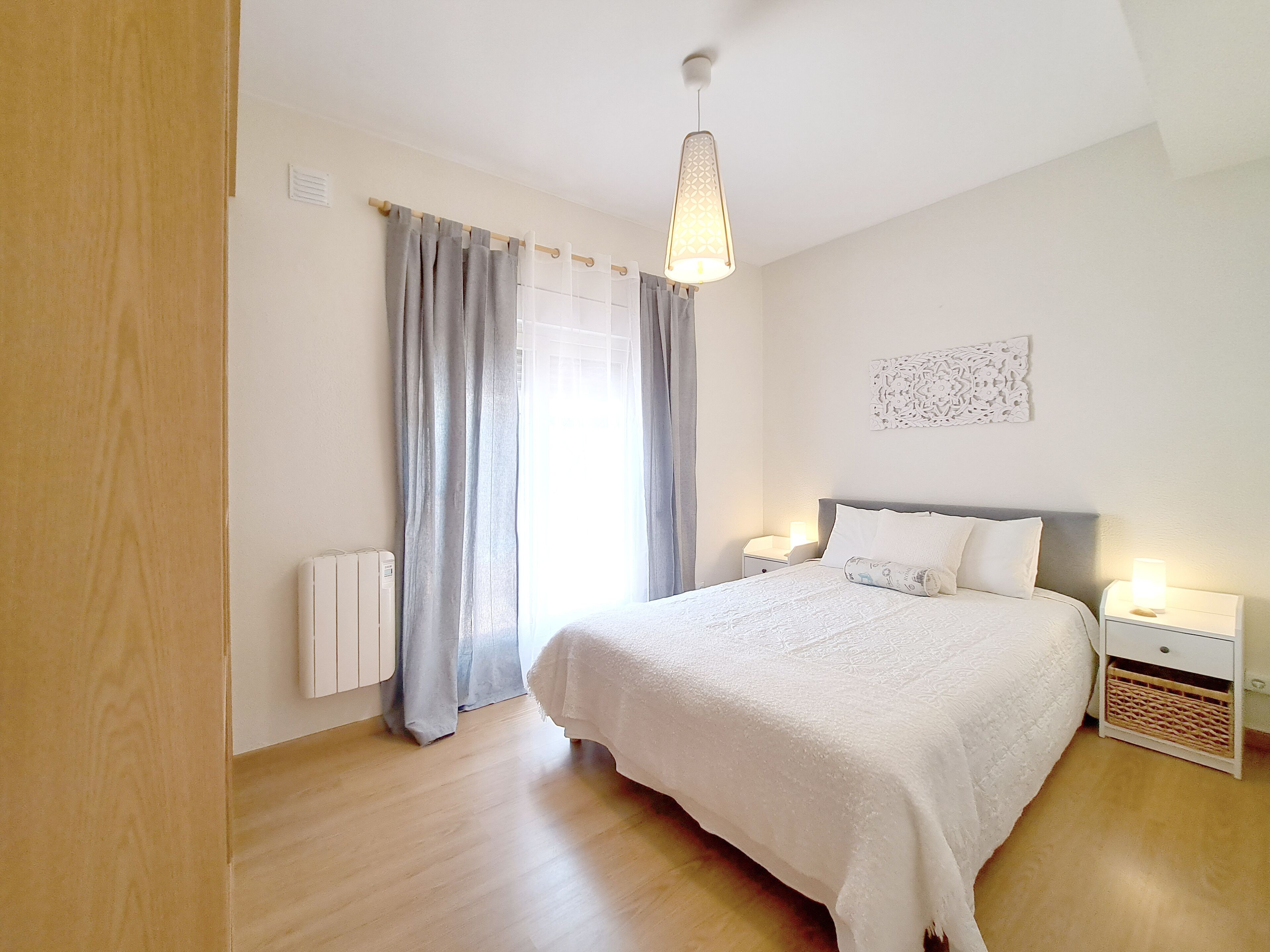 Un dormitorio con cama, mesitas de noche, un radiador, una ventana con cortinas y una lámpara colgante.