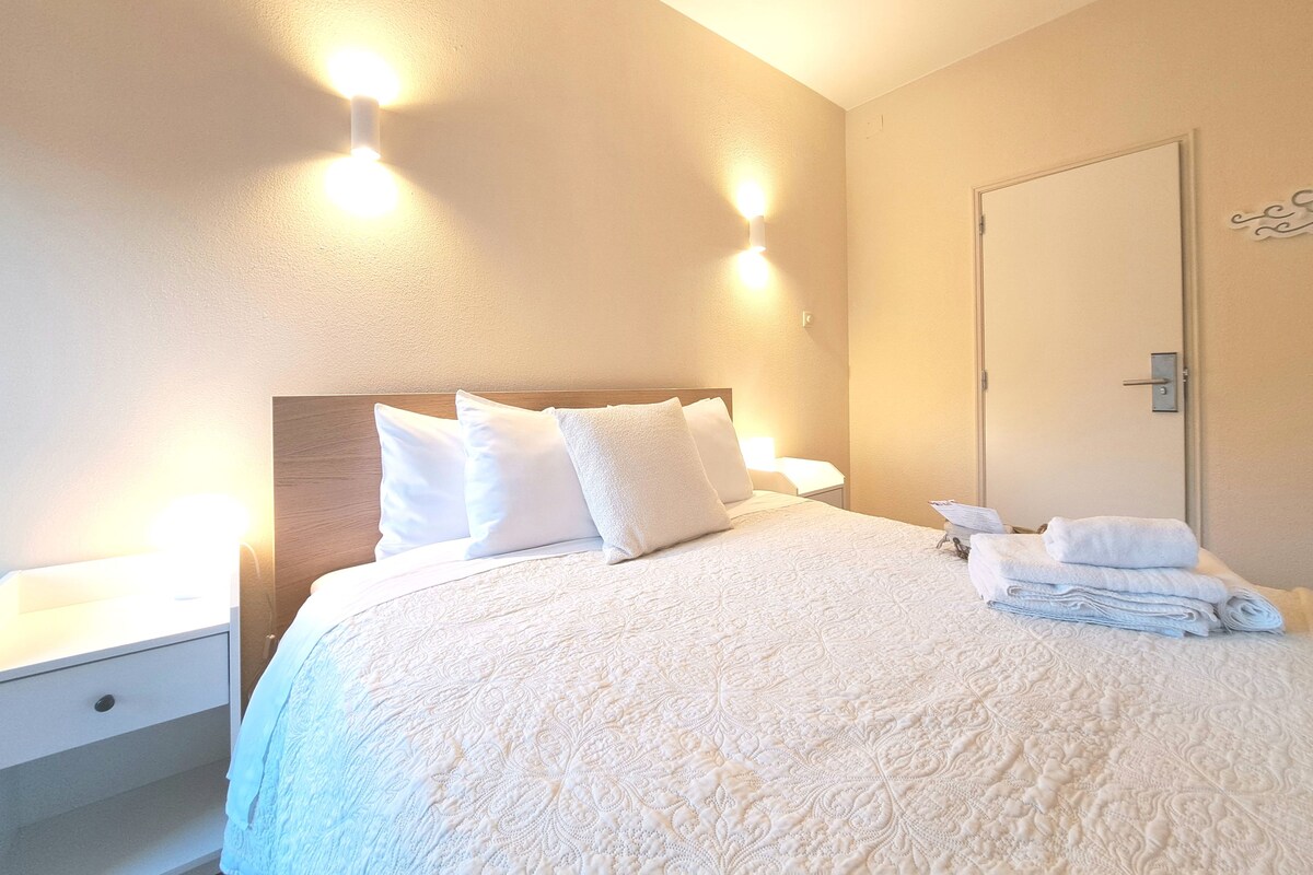 Een keurig opgemaakt bed met witte lakens, een nachtkastje met een lamp, en een deur met een handgreep.