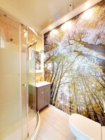A bathroom with a glass shower enclosure, a sink with a mirror, and a toilet. A mural of trees is on the wall.