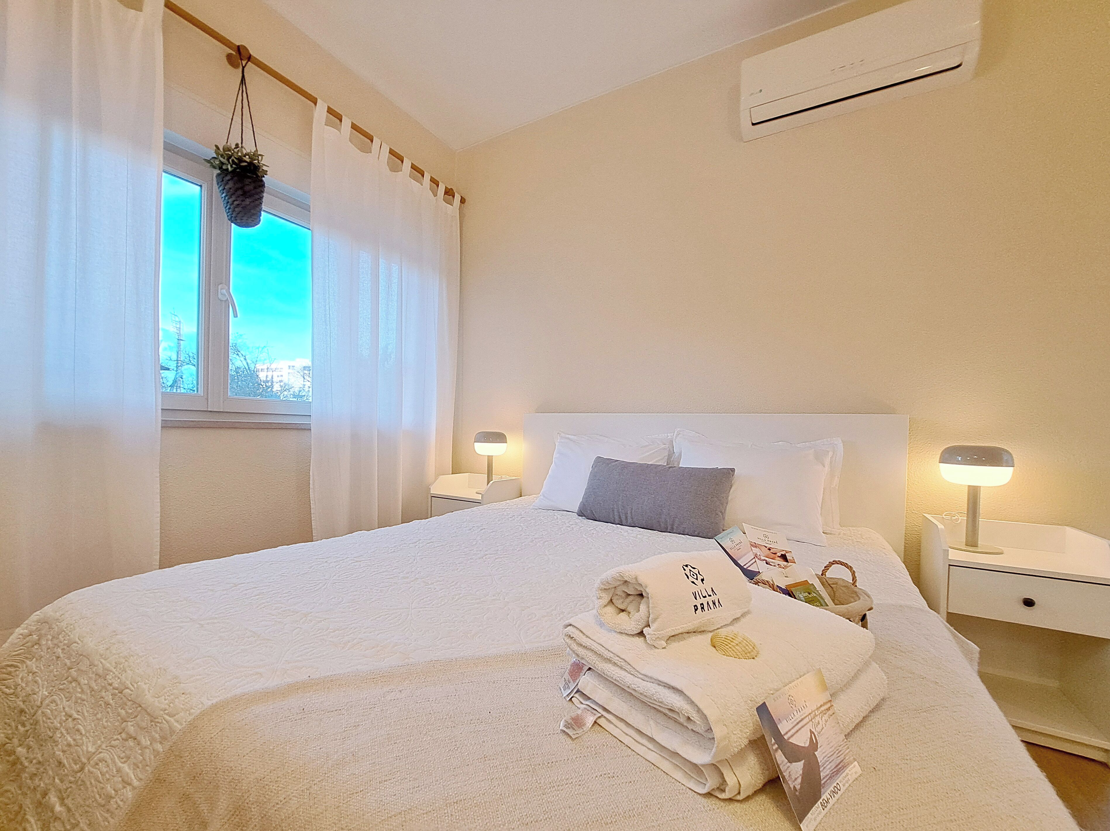 Uma cama bem arrumada com toalha dobrada e um livro, abajur de cabeceira e janela com cortinas.