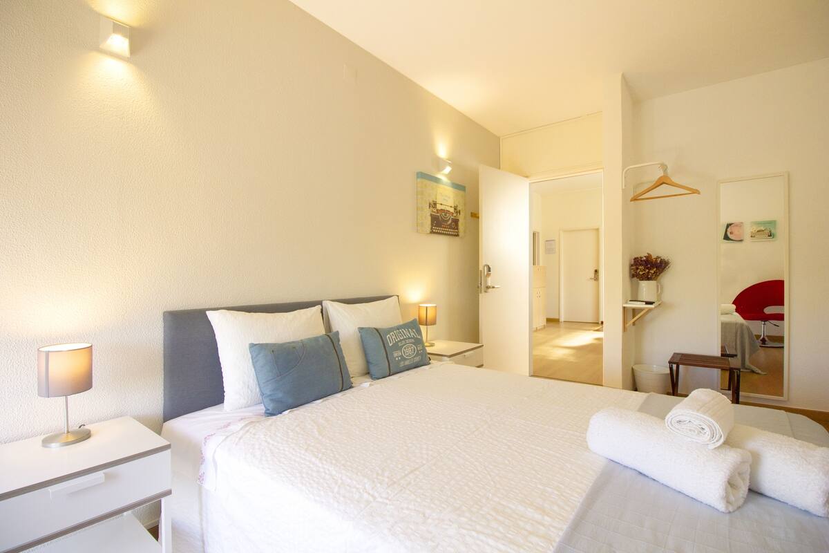 Ein Hotelzimmer mit einem ordentlich bezogenen Bett, Nachttischen, einem Spiegel und einer Tür zu einem weiteren Raum.