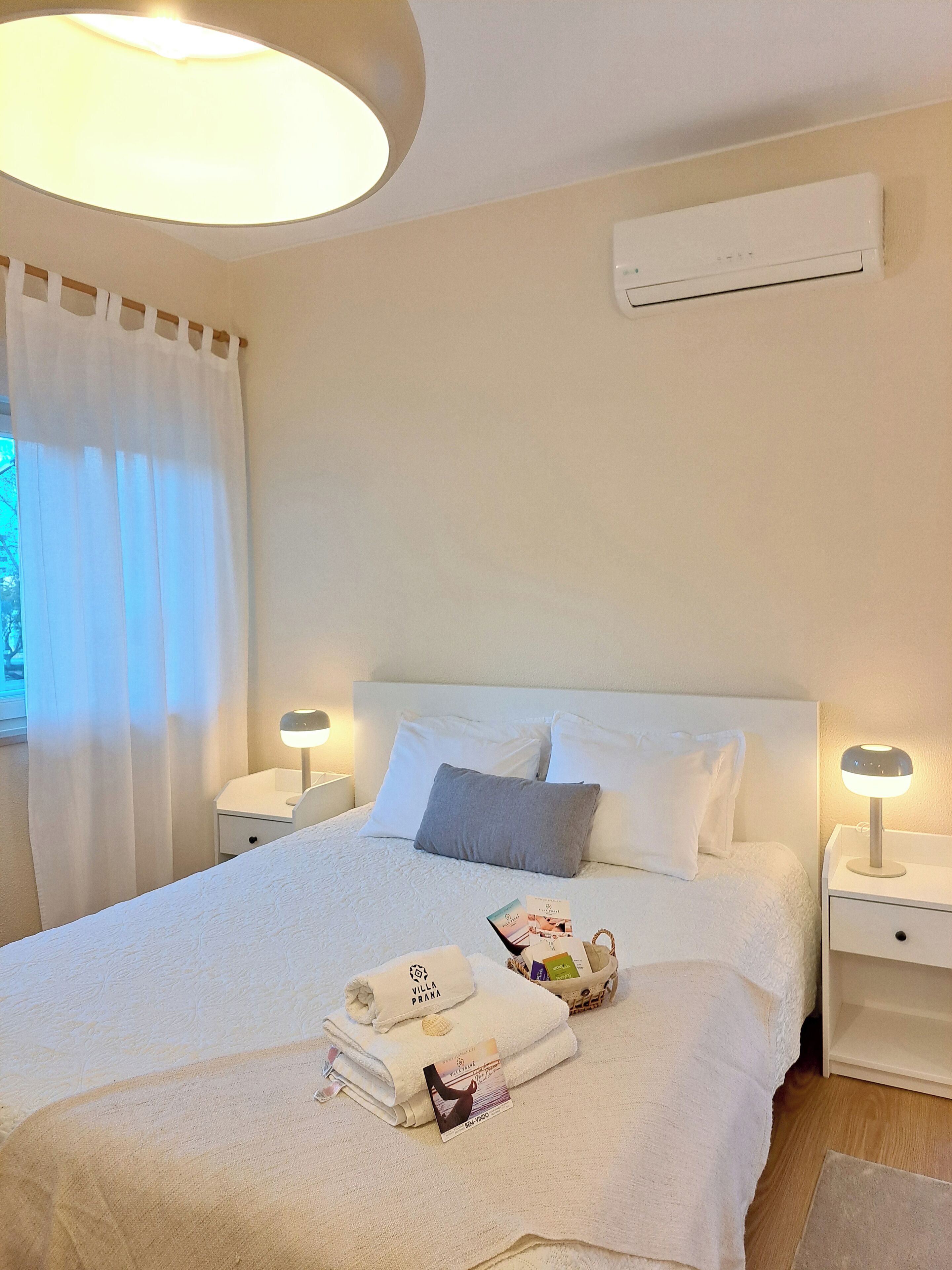 A neatly made bed with white linens, two bedside lamps, a white air conditioning unit, and a window with white curtains.