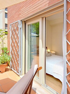 Une chambre avec un lit, une table de chevet et une lampe. Une porte-fenêtre coulissante donne sur un espace extérieur aménagé avec une chaise et des plantes en pot.
