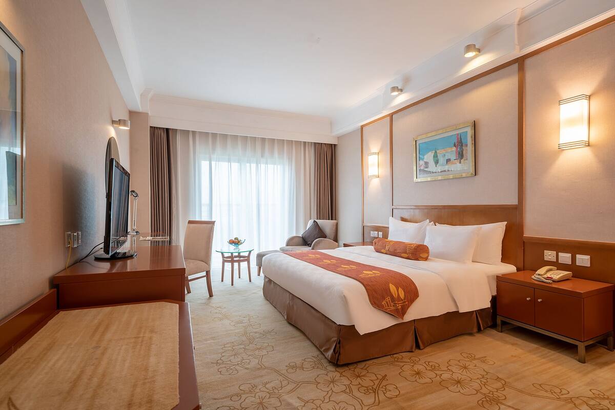 A hotel room with a large bed, a desk with a television, a small table with a chair, and a nightstand with a phone.