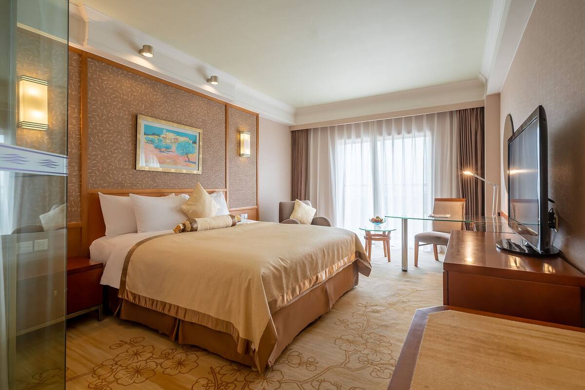 A hotel room with a large bed, a desk with a TV, a chair, a small table, and a painting on the wall.
