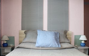 Ein Schlafzimmer mit einem Bett, zwei Nachttischen, jeweils einer Lampe und einem Fenster mit Jalousien.