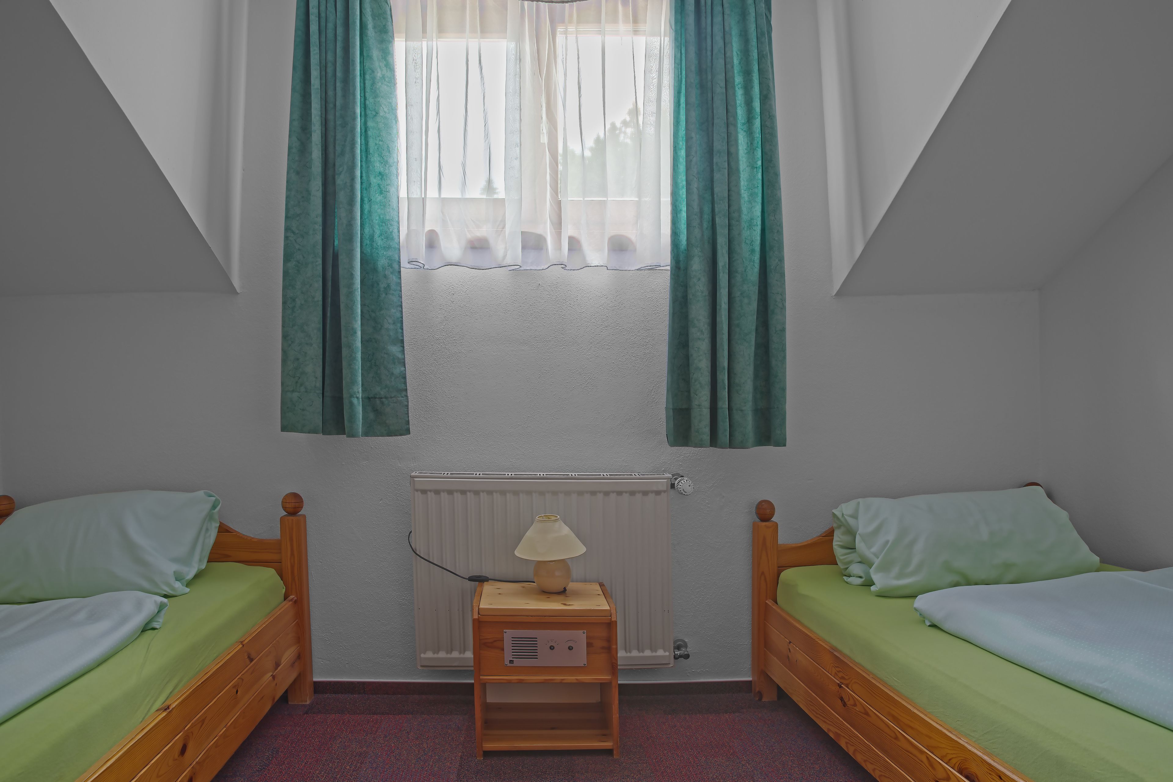 Une petite chambre confortable avec deux lits simples, une table de chevet avec une lampe, un radiateur et une fenêtre avec des rideaux.