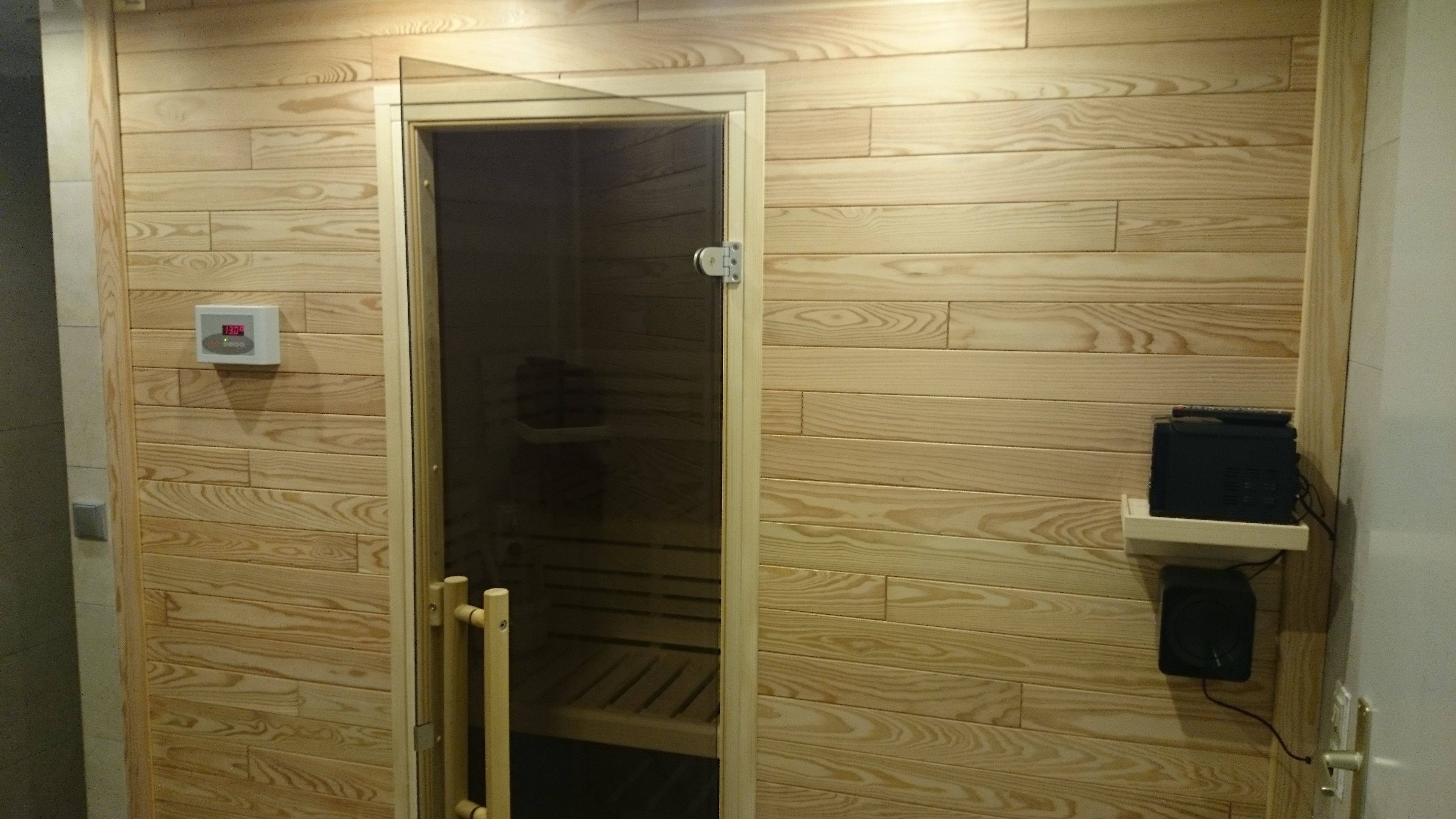 A sauna room with wooden walls, a glass door, a control panel, and a shelf with electronic devices.