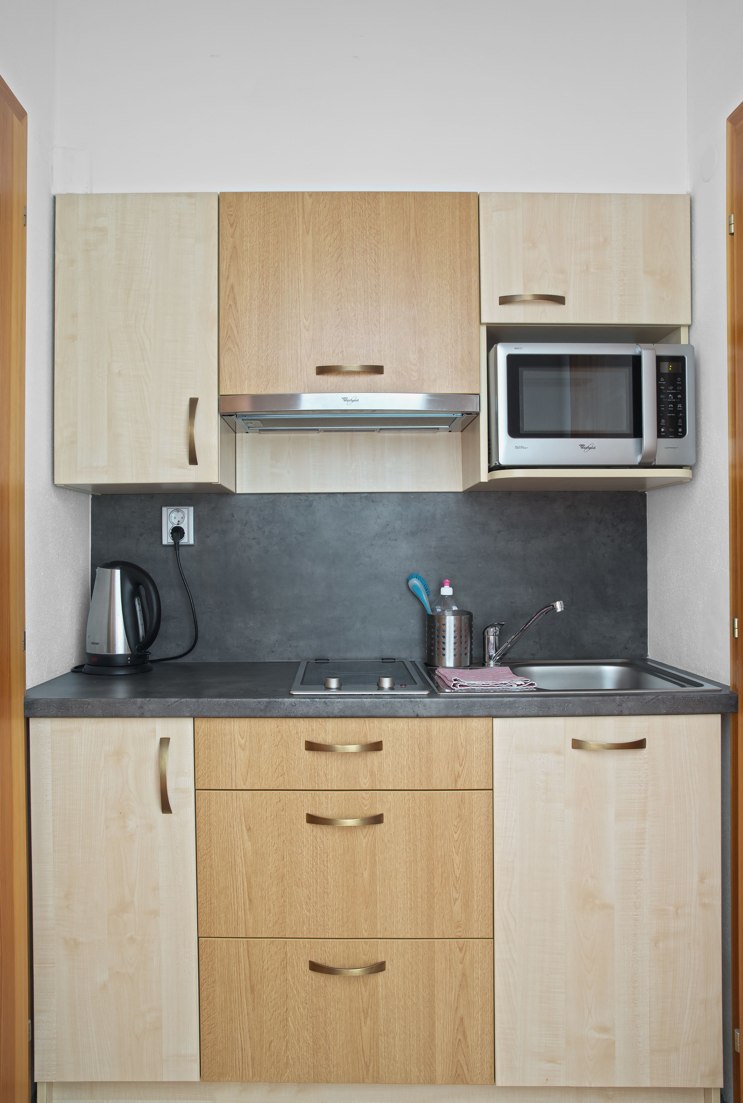 A compact kitchen with wooden cabinets, a built-in microwave, a kettle, and a sink with modern faucets.