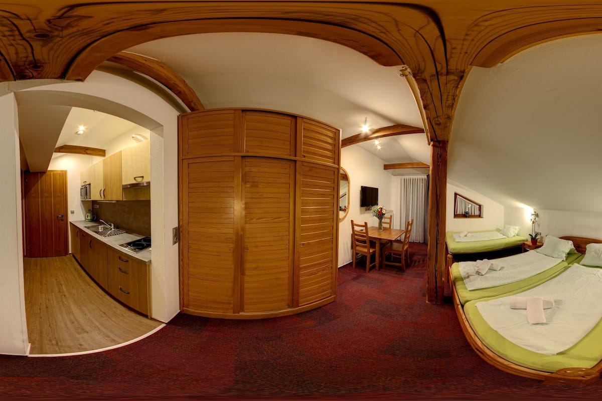 A room with a curved wooden ceiling, a bed with green bedding, a wooden wardrobe, a dining table with chairs, and a small bedside table with a lamp.