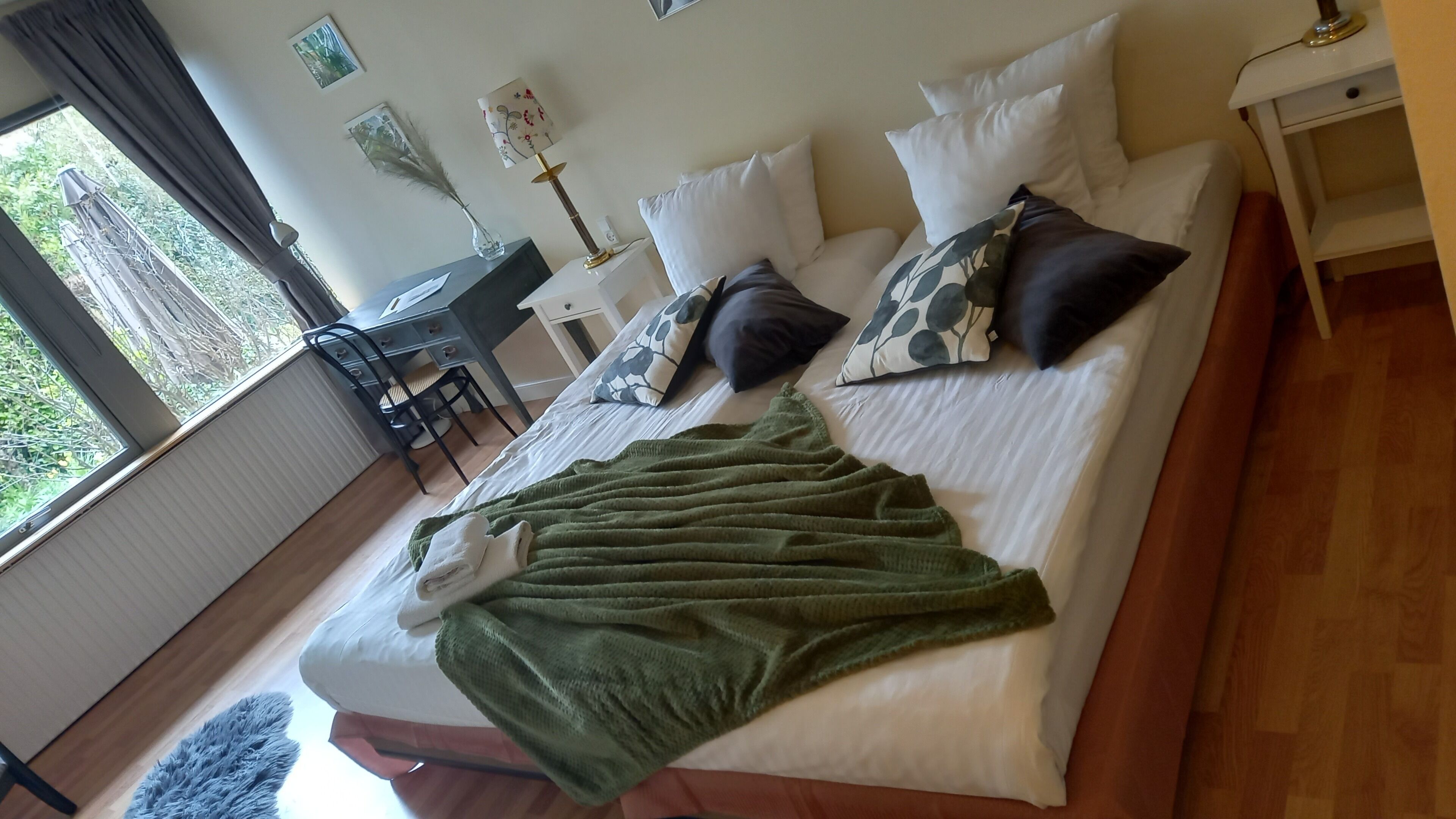 Una cama bien hecha con una manta verde, almohadas blancas y un escritorio con una silla en una habitación con un ventanal grande.
