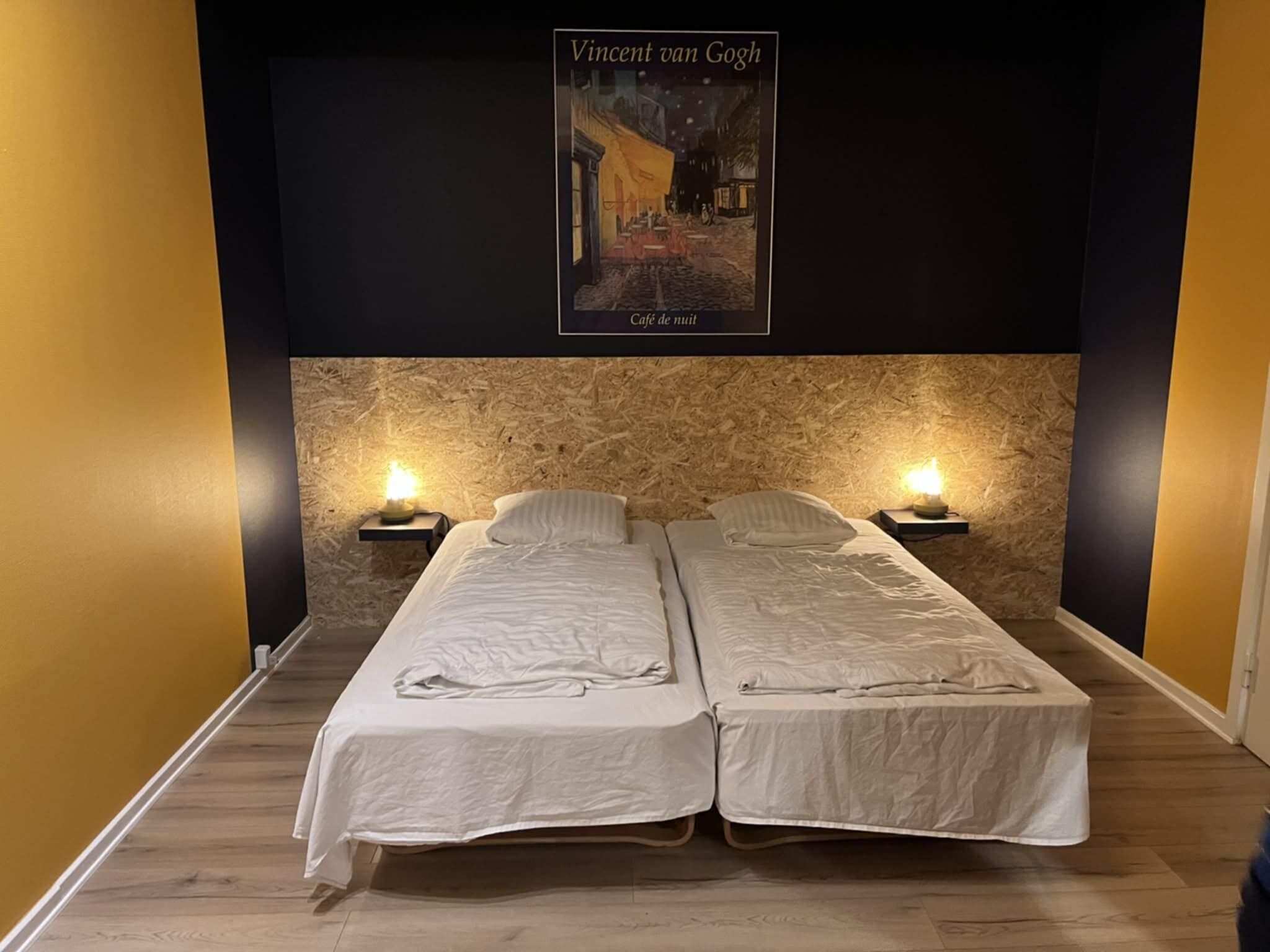 Una habitación con dos camas individuales, una pared de madera texturizada y un cartel del cuadro 'Café de Nuit' de Vincent van Gogh en la pared.