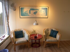 A room with two wicker chairs, a small round table with a potted plant, a framed artwork on the wall, and a window with a curtain. - Klintholm Bed & Breakfast (Borre)