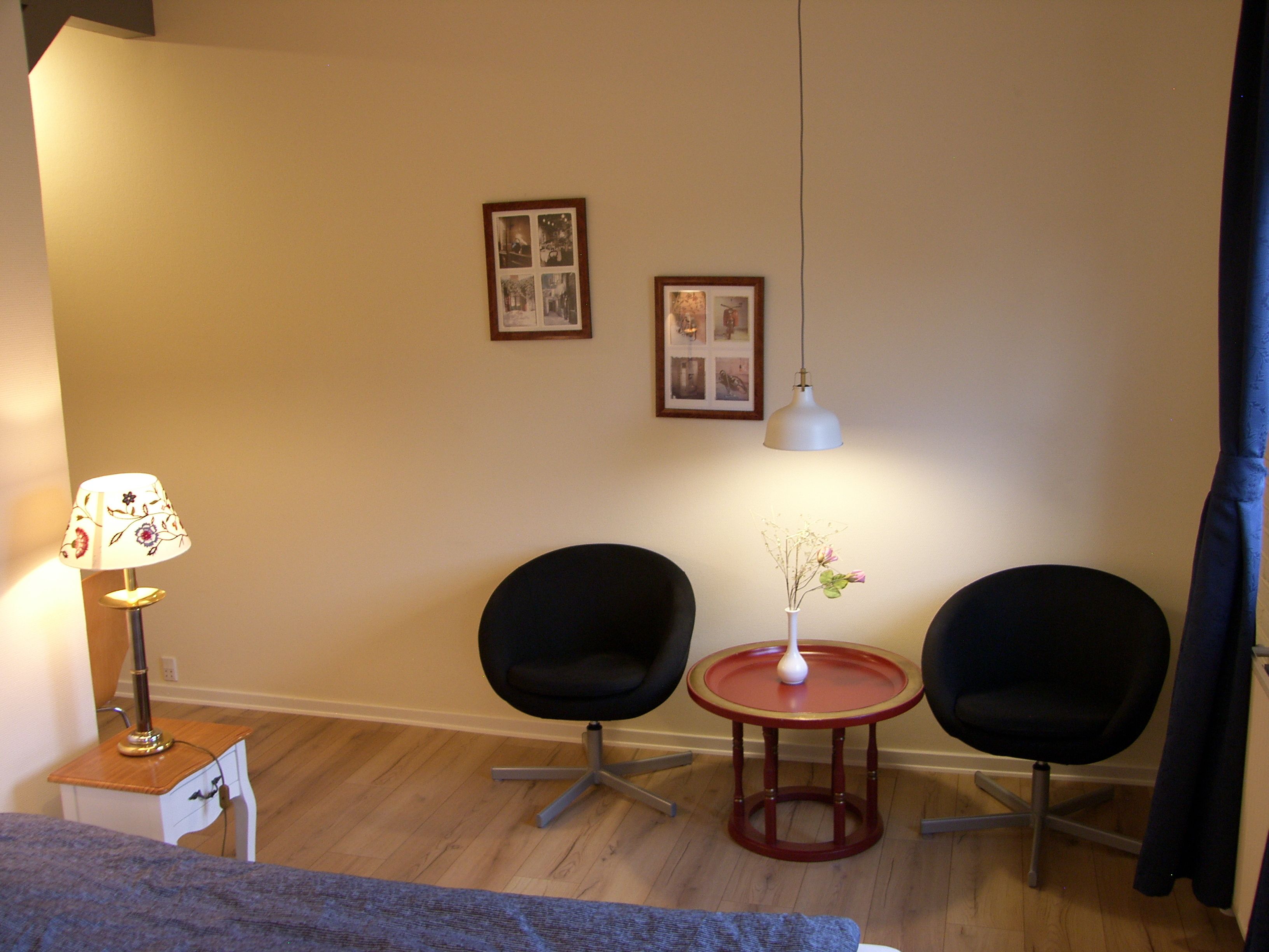 Una habitación pequeña y acogedora con dos sillas negras, una mesita con un florero, una lámpara y dos cuadros en la pared.