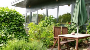 Un patio al aire libre con una mesa y sillas de madera, rodeado de abundante vegetación y una casa al fondo.