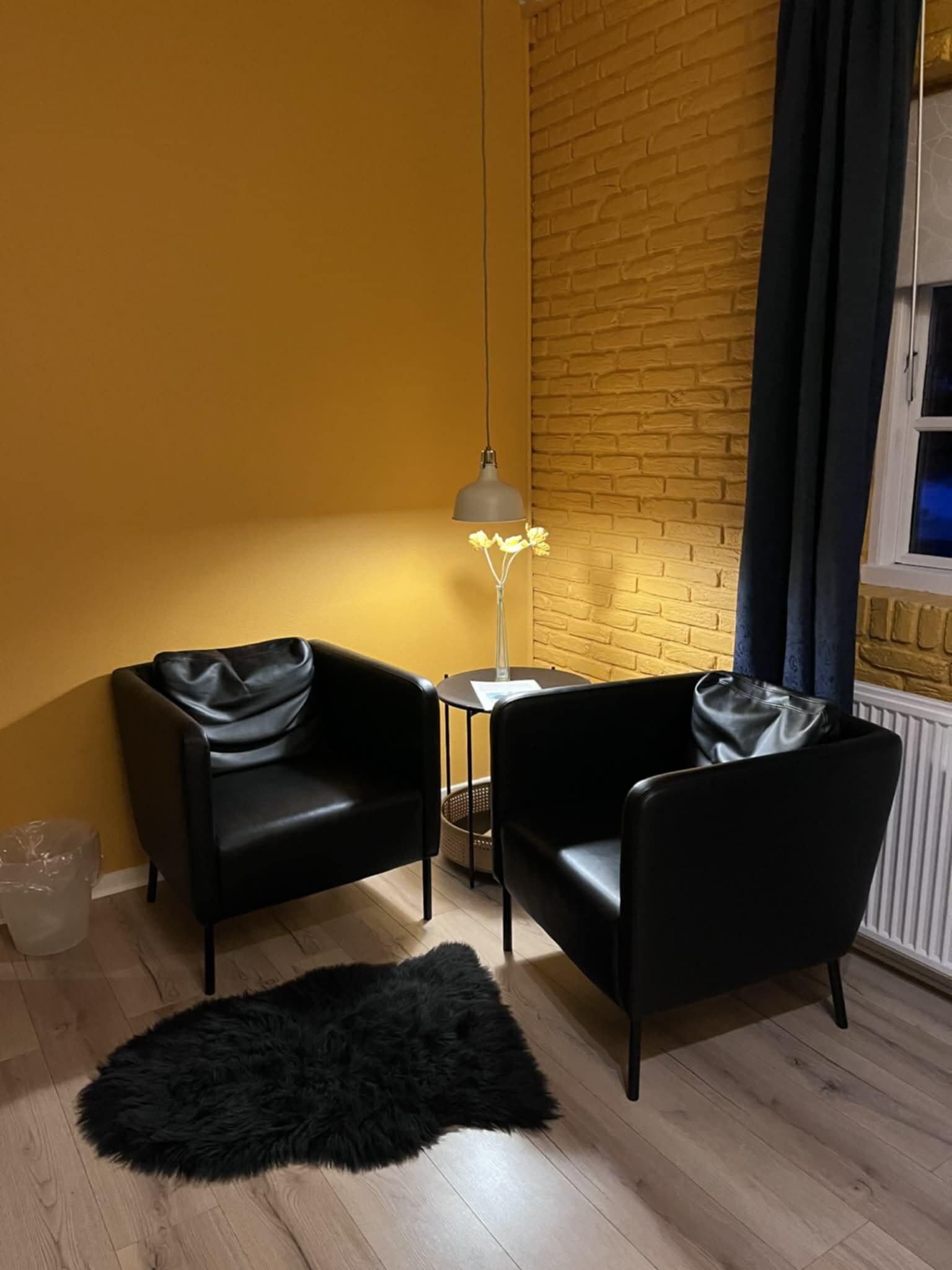 A room with two black leather chairs, a small round table, a lamp, a black furry rug, and a brick wall.