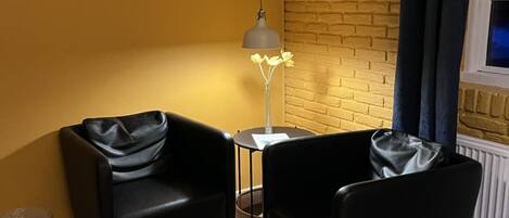 A room with two black leather chairs, a small round table, a lamp, a black furry rug, and a brick wall.