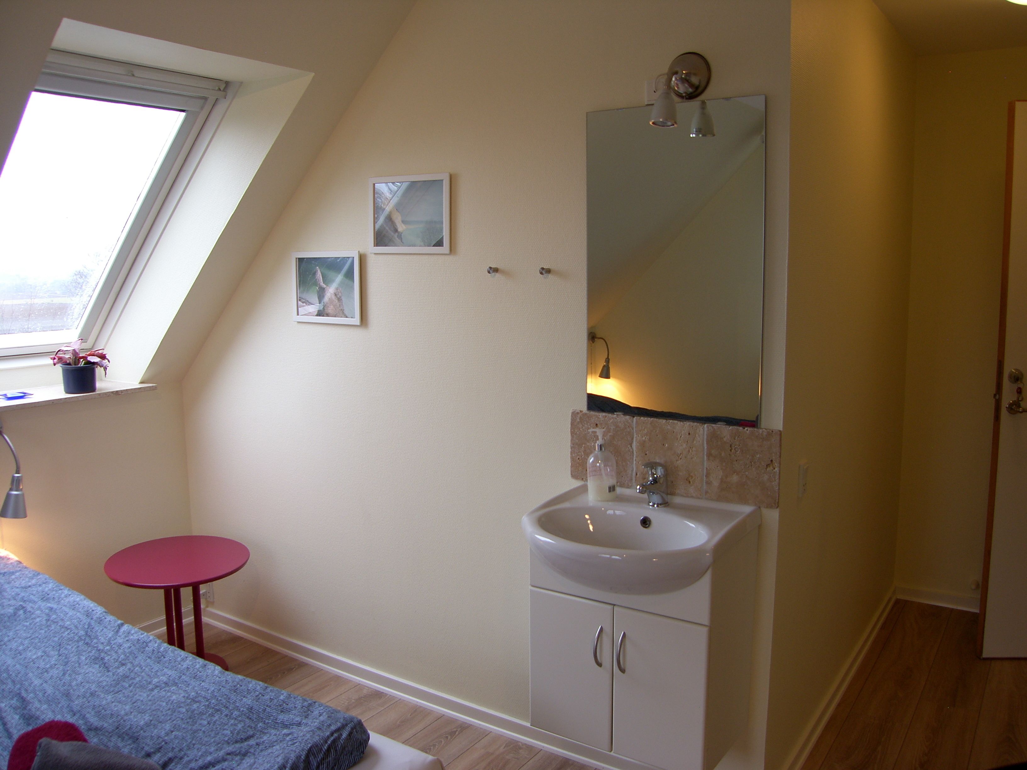Une chambre avec un puits de lumière, une table de chevet, un tabouret rouge, un lit, une salle de bain avec un lavabo et un miroir, et une porte.